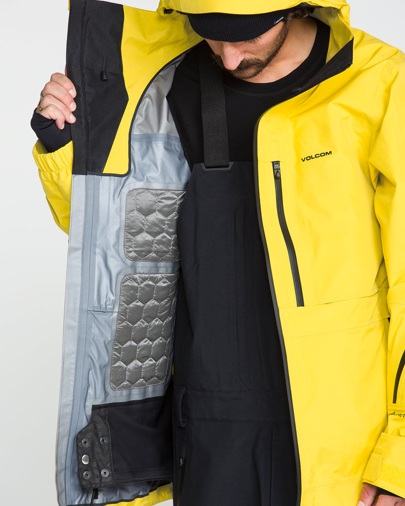 Wearing the Volcom Men’s TDS® Inf Gore-Tex Jacket in yellow, a person opens the jacket to reveal its detailed interior, including a quilted gray panel and black inner lining. The partially visible model showcases the jacket’s premium GORE-TEX construction, emphasizing advanced materials and craftsmanship.