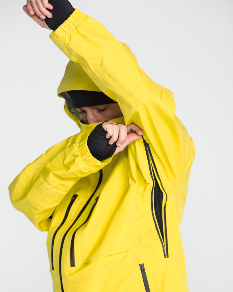 A person wearing the Volcom Men’s TDS® Inf Gore-Tex Jacket in yellow, hood up and partially concealing their face with their arm and sleeve, stands against a plain light background.