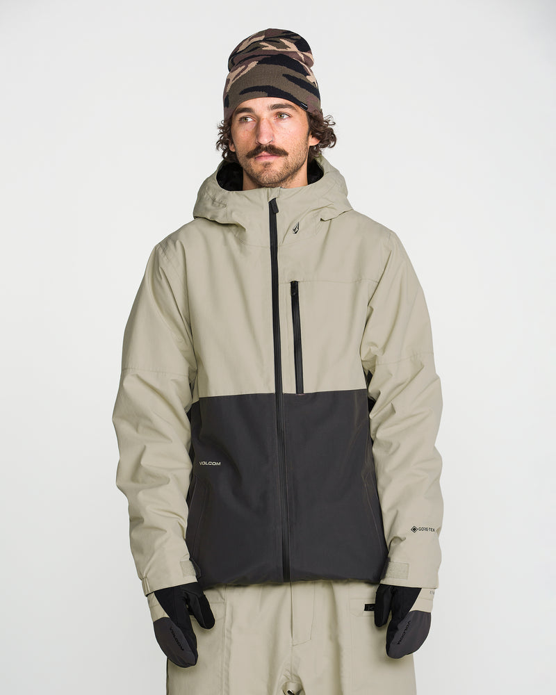 A person wearing the Volcom Men’s L Insulated Gore-Tex Jacket in beige and black, paired with beige pants, black gloves, and a patterned beanie stands against a plain white background—ready for waterproof protection in any weather.