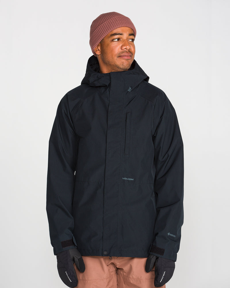 A person wearing the Volcom Men’s Dua Insulated Gore-Tex Jacket in black, paired with a light brown beanie, black gloves, and pinkish-brown pants stands against a plain light background, looking slightly to their left with a subtle smile.