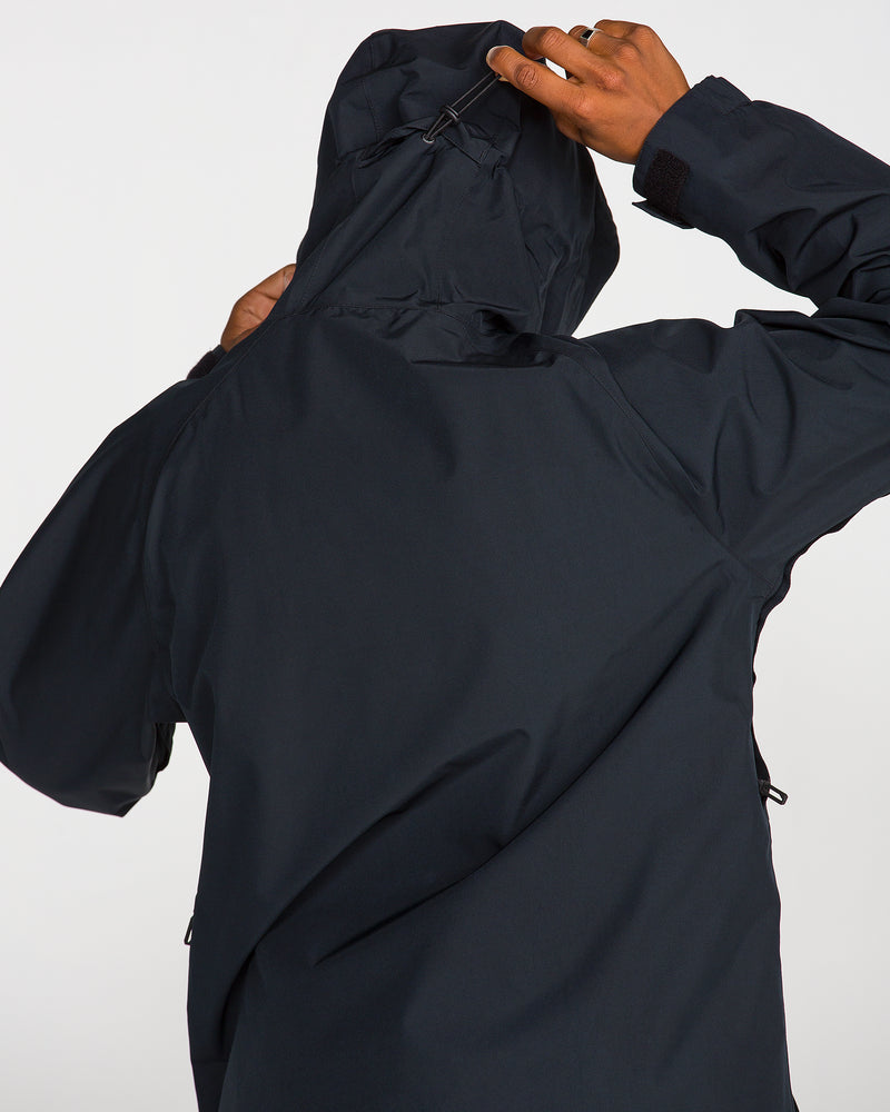 A person wearing the Volcom Men’s Dua Insulated Gore-Tex Jacket is seen from behind, adjusting the hood with one hand. The jacket, featuring a matte finish and long sleeves, offers reliable weather protection. The background is plain and light-colored.