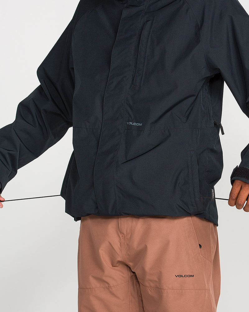 A person wearing the Volcom Men’s Dua Insulated GORE-TEX Jacket in black and light brown Volcom pants tightens the jacket’s hem drawstring. Only the torso and part of the arms are visible against a plain background, showcasing the sleek, waterproof design of the jacket.