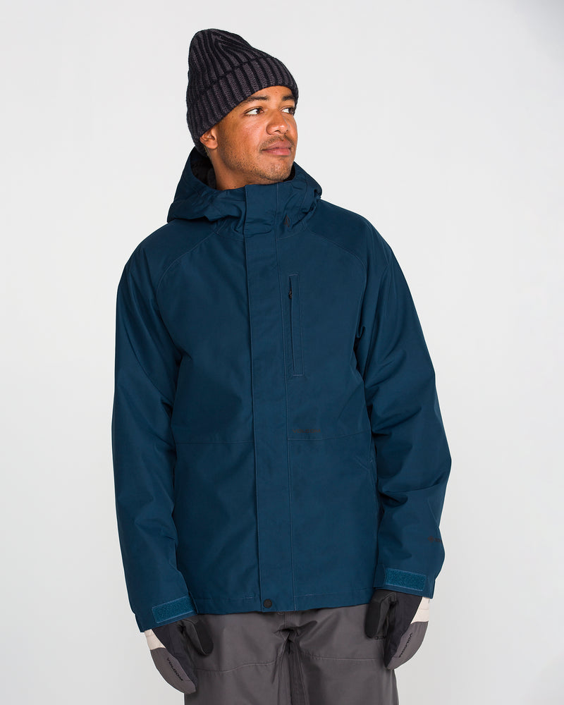 A person wearing the Volcom Men’s Dua Insulated Gore-Tex Jacket in dark blue, paired with gray gloves, gray pants, and a black knit beanie, stands against a plain light background and looks slightly to the side.