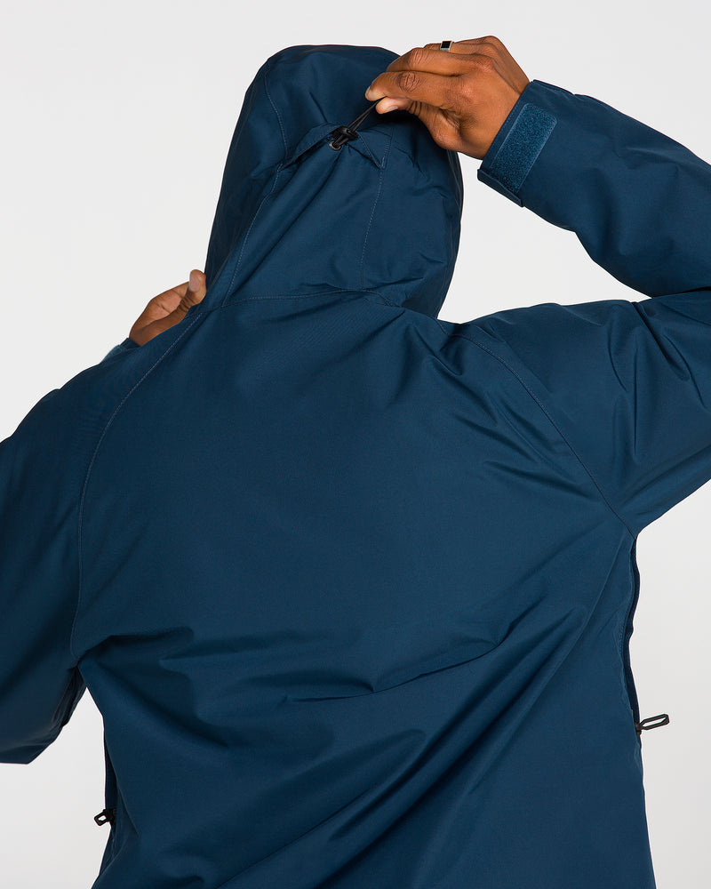 A person wearing the Volcom Men’s Dua Insulated Gore-Tex Jacket in dark blue adjusts the hood by pulling a drawstring at the back. The photo is taken from behind, highlighting their upper body and hand.
