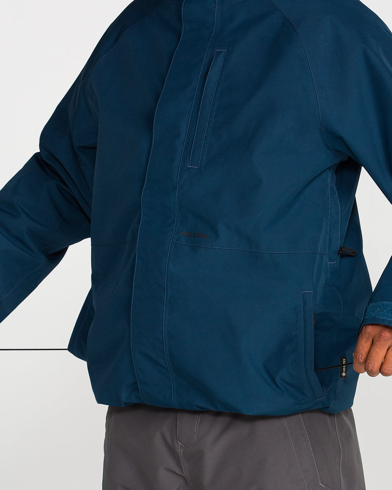A person wearing the Volcom Men’s Dua Insulated Gore-Tex Jacket in blue and gray pants stands with one arm outstretched, displaying the front and side of the waterproof insulated ski jacket. The image is cropped at the neck and mid-thigh.