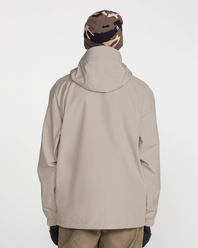A person is standing with their back to the camera against a plain white background, wearing the Volcom Men’s Dua Insulated Gore-Tex Jacket in beige, featuring recycled insulation, along with brown pants, black gloves, and a brown camo beanie.