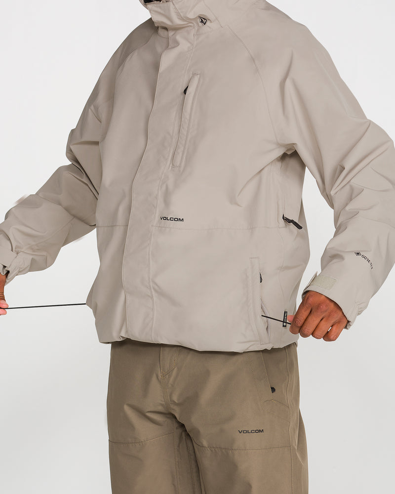 A person wearing the Volcom Men’s Dua Insulated GORE-TEX Jacket in light beige with matching pants tightens the jacket’s waist drawstrings. The face is partially out of frame. This waterproof ski jacket features zippered pockets, a high collar, and recycled insulation for added warmth.