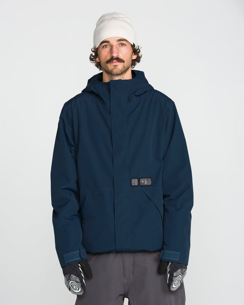 A person with a mustache and curly hair wears a white beanie, gray pants, patterned gloves, and the Volcom Men’s Double Insulated Jacket featuring Zip Tech in navy blue, standing against a plain white background.