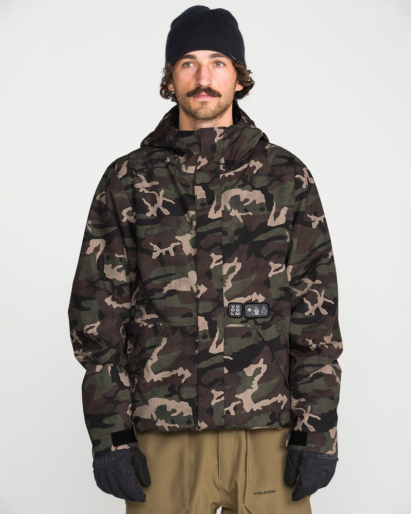 A man with a mustache wears a black beanie, tan pants, and the Volcom Men’s Double Insulated Jacket—a camo-patterned technical outerwear jacket featuring 10K/10K V-Science technology and recycled insulation—standing against a plain white background with his gloved hands at his sides.