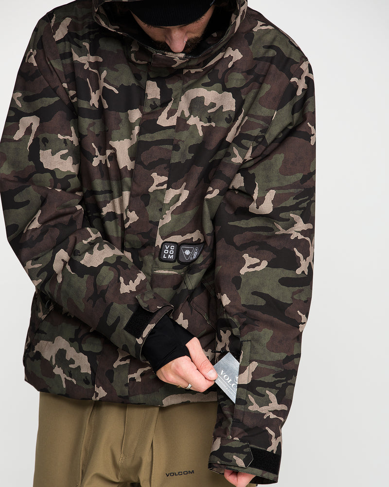 A person wearing the Volcom Men’s Double Insulated Jacket—a camo technical outerwear piece with a hood, Velcro wrist straps, and recycled insulation—pulls a tag reading “Volcom” from the sleeve while dressed in tan pants. The individual’s face is not visible.