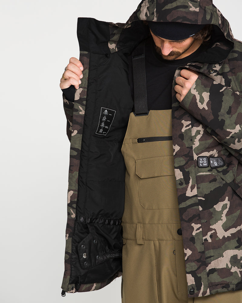 Wearing the Volcom Men’s Double Insulated Jacket, a person in brown overalls opens the camouflage technical outerwear to show its black inner lining and a label featuring symbols that emphasize its 10K/10K V-Science performance. With their face partly concealed by the jacket’s hood and a beanie, they highlight both recycled warmth and functional design.