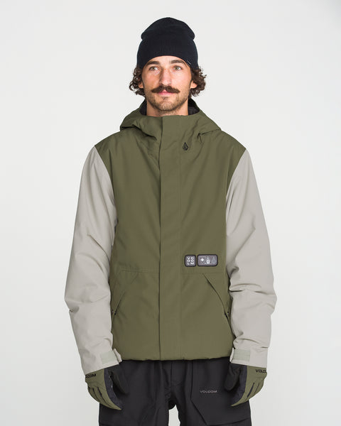 A man with a mustache wears a black beanie, black gloves, and the Volcom Men’s Double Insulated Jacket—an olive green and gray technical outerwear piece featuring recycled warmth—as he stands facing forward against a plain white background.