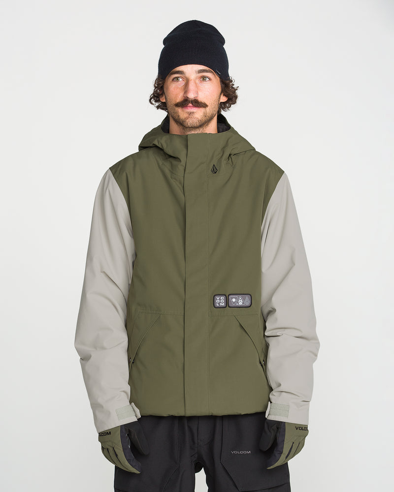 A man with a mustache wears a black beanie, black gloves, and the Volcom Men’s Double Insulated Jacket—an olive green and gray technical outerwear piece featuring recycled warmth—as he stands facing forward against a plain white background.