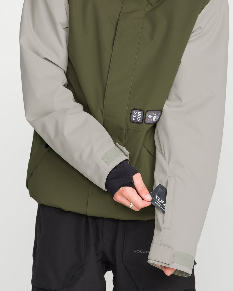 A person wearing the Volcom Men’s Double Insulated Jacket in olive green and light gray adjusts a black pocket on the left sleeve. Visible brand labels and innovative details, such as the Zip Tech jacket-to-pant interface for enhanced function, are featured on the jacket. The person’s face is not visible.