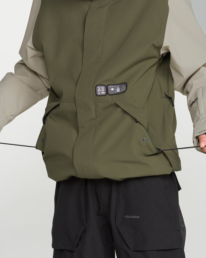 A person wears the Volcom Men’s Double Insulated Jacket—an olive green and beige technical outerwear piece crafted with recycled warmth materials, showcasing patches, visible pockets, and a drawstring waist—paired with black pants.