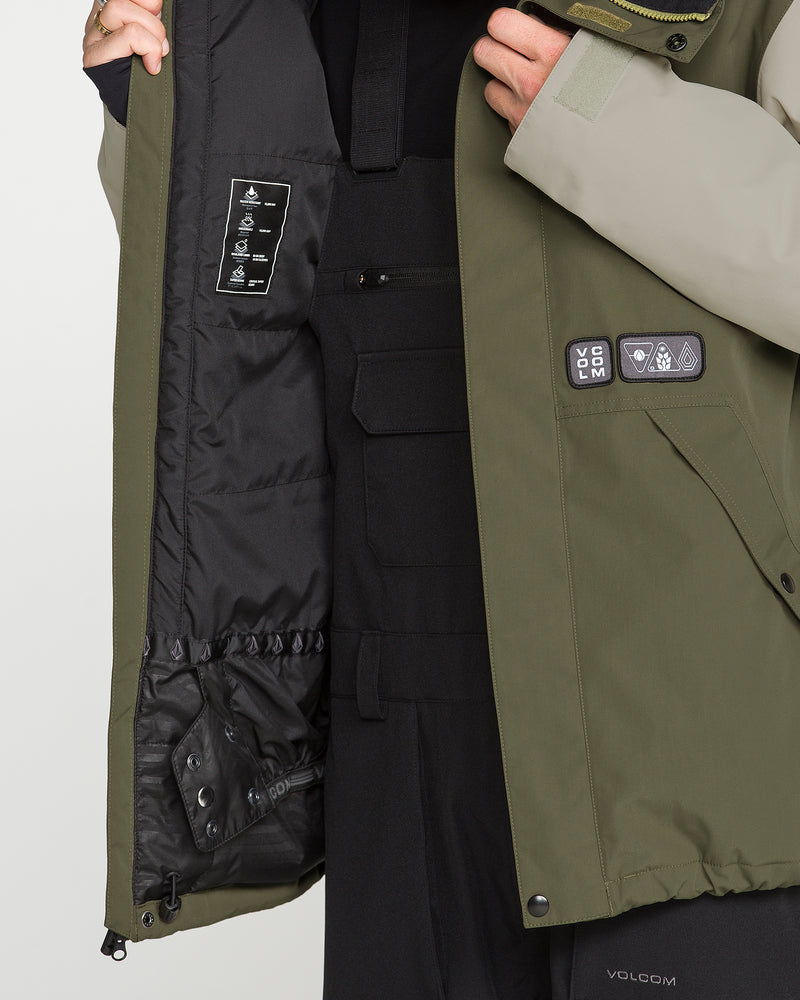 A person is shown wearing the Volcom Men’s Double Insulated Jacket in olive green, partially unzipped to reveal the black inner lining and layers beneath. Designed for recycled warmth, this technical outerwear features a patch with icons and a flap pocket. Only the torso and one arm are visible.