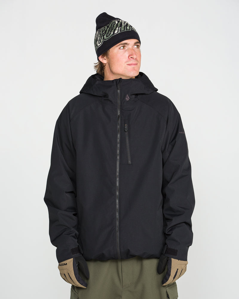 A man wearing the Volcom Men’s 2836 Insulated Jacket in black, paired with olive pants, tan gloves, and a black beanie with a white design, stands against a plain white background, looking slightly to the side.