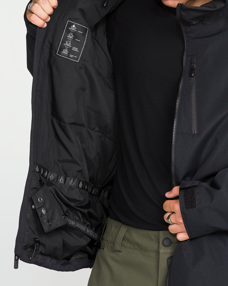 A man wearing olive green pants and a black shirt opens a Volcom Men’s 2836 Insulated Jacket in black to display its inner label, pockets, and elastic waistband details; only his torso and part of his face are visible.