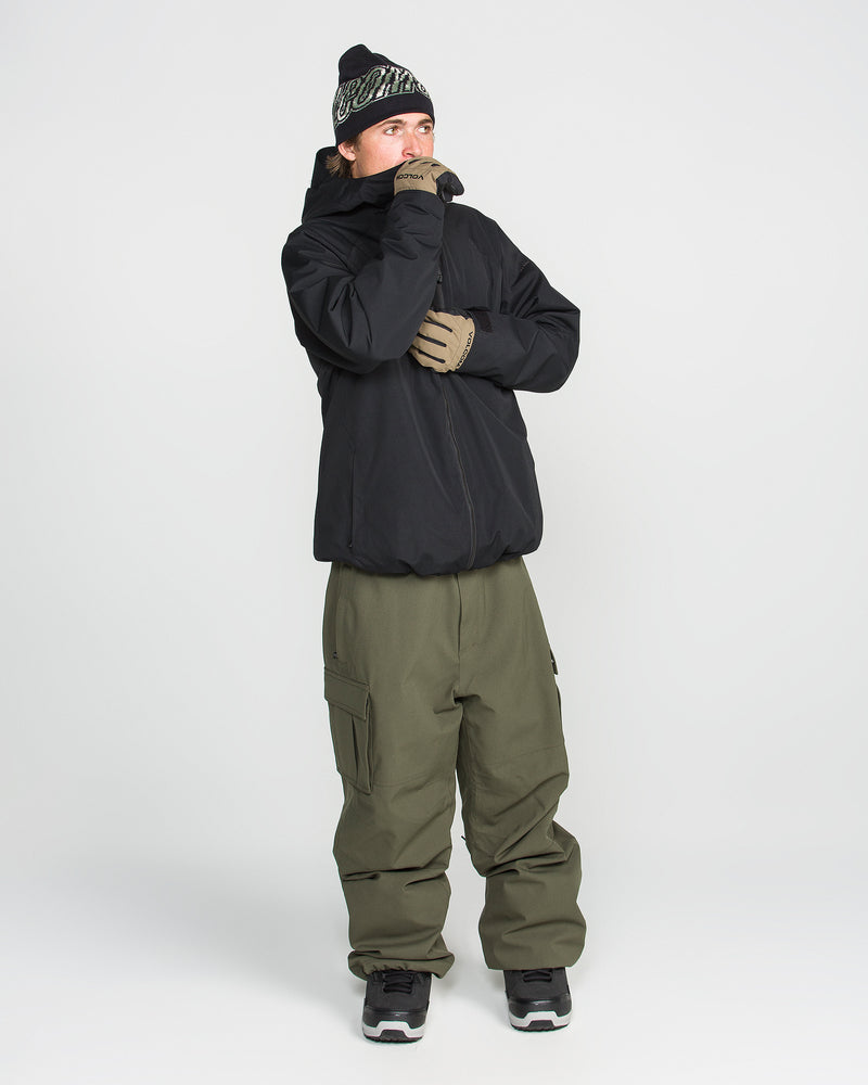 A person wearing the Volcom Men’s 2836 Insulated Jacket, along with olive green cargo pants, a black beanie, gloves, and sneakers, stands against a plain white background looking to the side with their hand near their mouth.