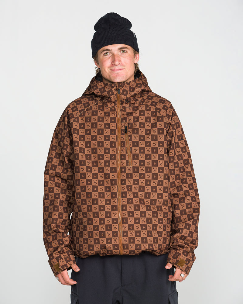 A person wearing the Volcom Men’s 2836 Insulated Jacket in a brown pattern, which features V-Science 2-Layer technology and 10K waterproof protection, along with a hood and a black beanie, stands against a plain white background, looking at the camera and smiling slightly.