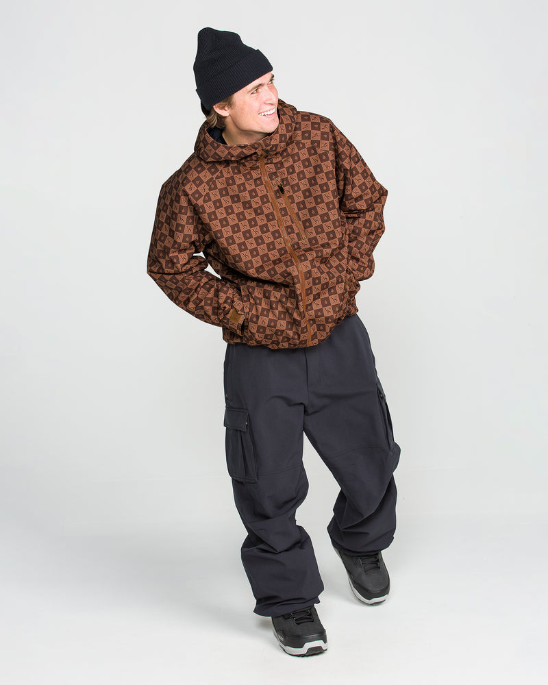 Smiling person wearing a black beanie, the Volcom Men’s 2836 Insulated Jacket in a patterned brown color, loose black cargo pants, and black sneakers stands against a plain white background.