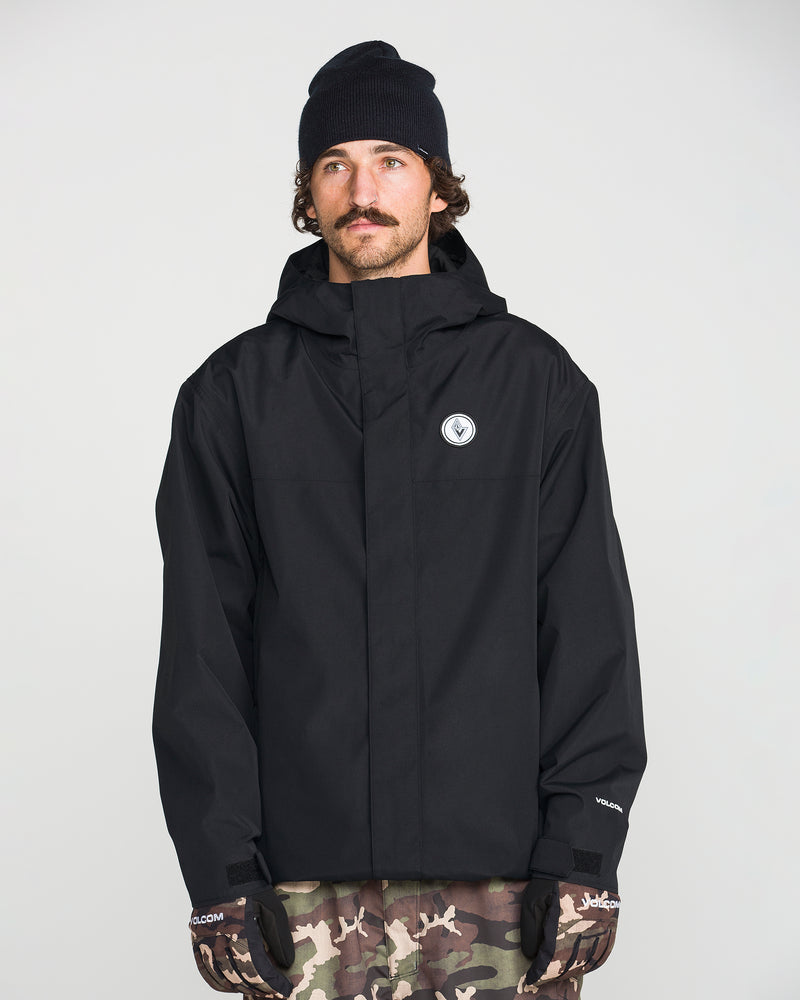 A man wearing the Volcom Men’s Buckthreeeighty Insulated Jacket made from recycled fabric, a black beanie, and camo pants stands facing forward with a neutral expression. He is also wearing black gloves and has a mustache. The background is plain and light-colored.