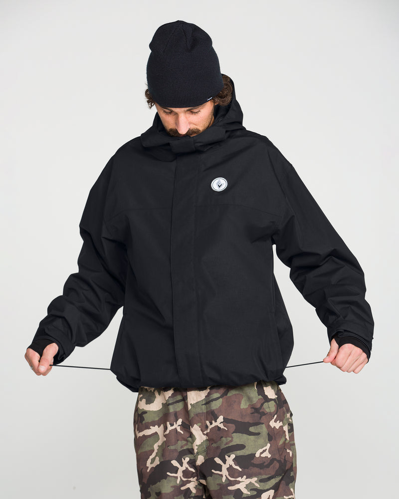 A person wearing the Volcom Men’s Buckthreeeighty Insulated Jacket in black and a black beanie adjusts the jacket’s hem drawstrings. They also have on camouflage pants and are standing against a plain light background.