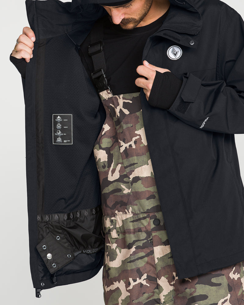 A person wearing the Volcom Men’s Buckthreeeighty Insulated Jacket in black and camouflage overalls opens the jacket to reveal its inner lining and a care label, with only their torso and part of their face visible.