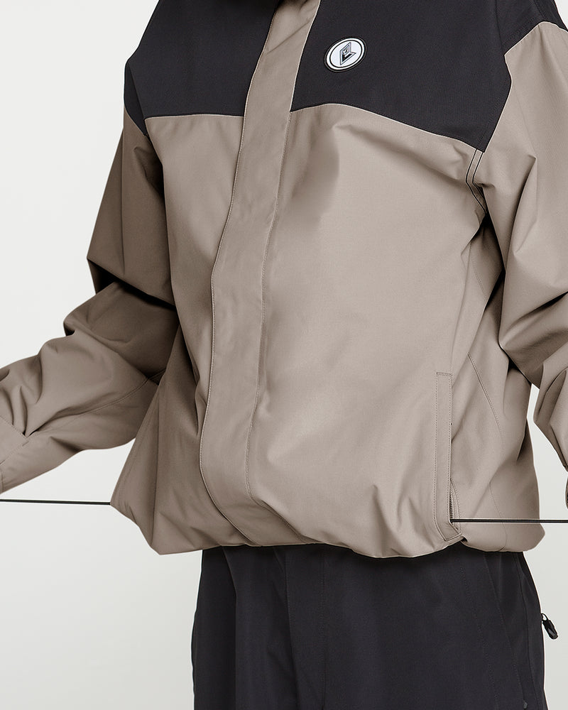 A person is wearing the Volcom Men’s Buckthreeeighty Insulated Jacket in taupe and black, featuring a round logo on the chest; only their torso and part of their arms are visible as they stand against a plain background.
