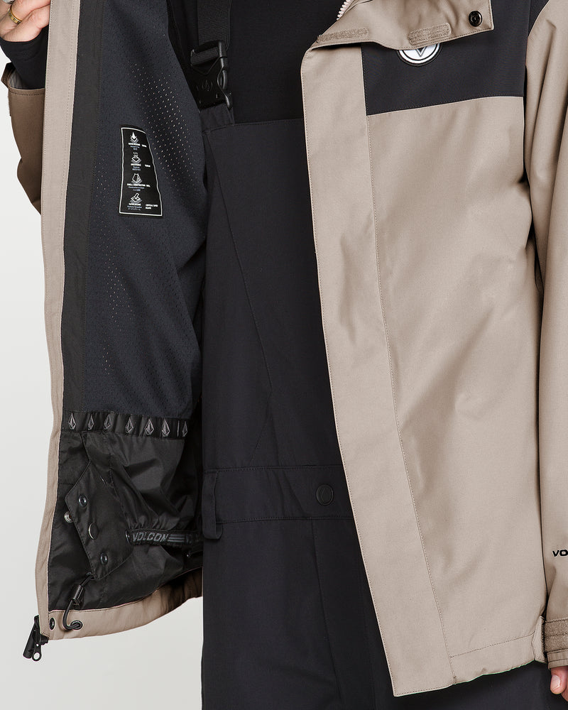 A person opens the Volcom Men’s Buckthreeeighty Insulated Jacket in tan and black, revealing its interior lining and the care instructions label. Made from recycled fabric, this jacket combines stylish design with eco-friendly materials. Underneath, the person is dressed in black clothing.