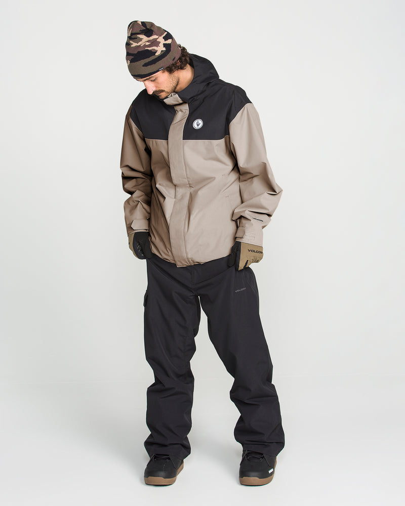 A person wearing the Volcom Men’s Buckthreeeighty Insulated Jacket in tan and black—crafted from recycled fabric with 10K waterproof insulation—along with black pants, gloves, a camouflage beanie, and black shoes stands looking down against a plain white background.