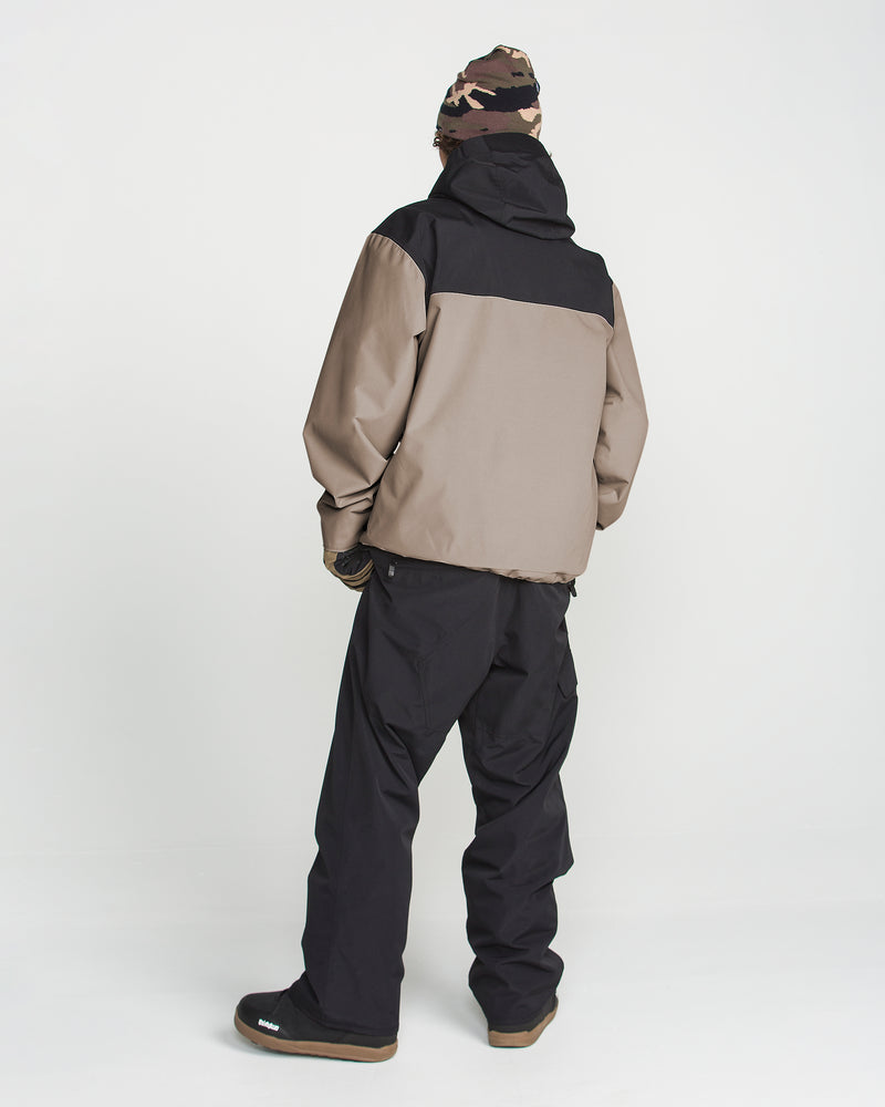 A person stands with their back to the camera against a plain white background, wearing the Volcom Men’s Buckthreeeighty Insulated Jacket in beige and black—crafted from recycled fabric—paired with black pants, black shoes, gloves, and a camouflage beanie.