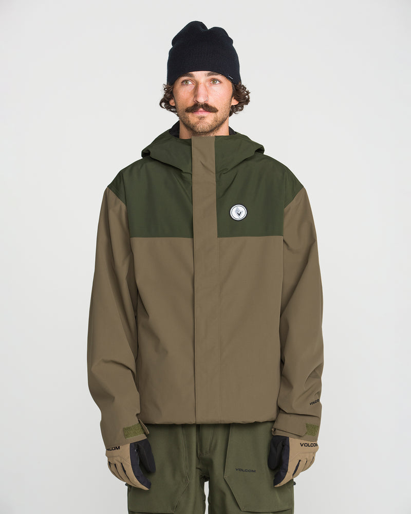A man stands facing forward against a plain white background, wearing the Volcom Men’s Buckthreeeighty Insulated Jacket in two-tone green and olive—crafted from recycled fabric—paired with olive pants, a black beanie, and gloves.