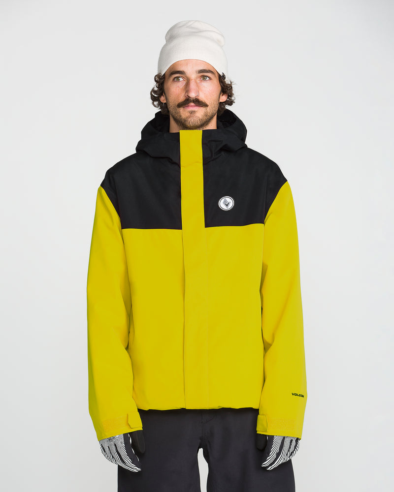 A man wearing the Volcom Men’s Buckthreeeighty Insulated Jacket—yellow and black and crafted from recycled fabric—stands against a plain white background, paired with a white beanie and patterned gloves, looking slightly to the side.