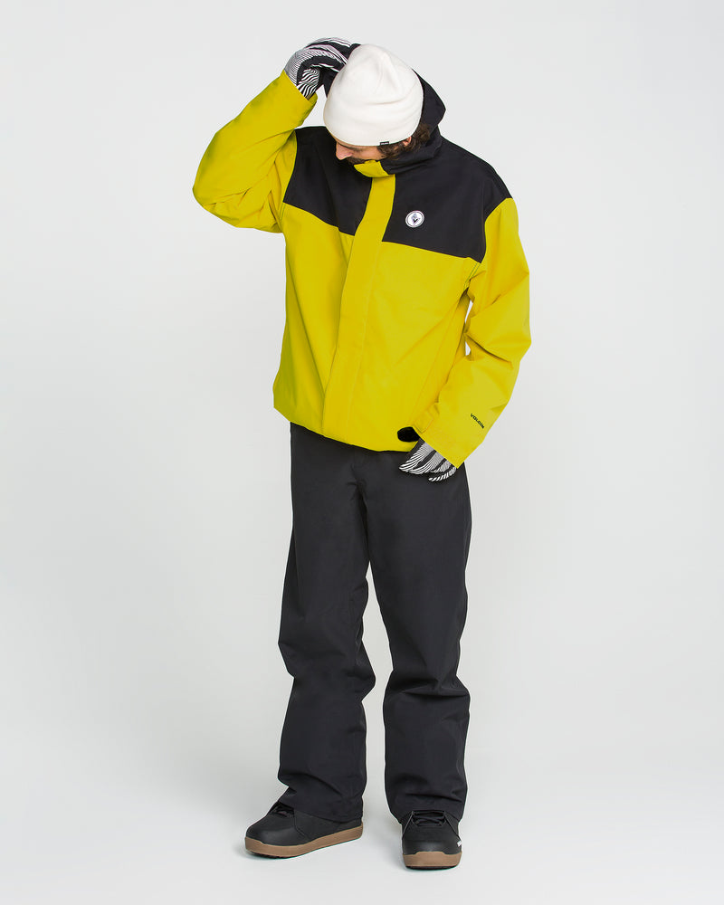 A person wearing the Volcom Men’s Buckthreeeighty Insulated Jacket in yellow and black, made from recycled fabric, along with black pants, striped gloves, a white beanie, and brown shoes stands against a plain white background, looking down with one hand on their head.