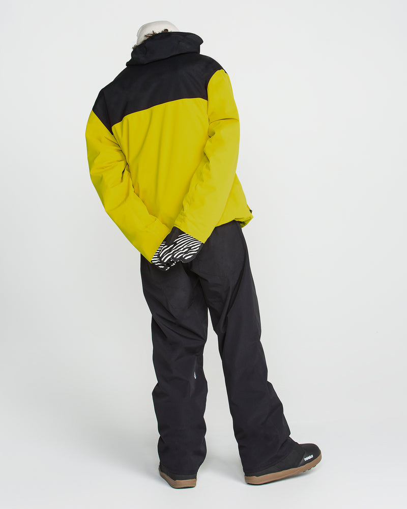 A person in the Volcom Men’s Buckthreeeighty Insulated Jacket, which features 10K protection, stands with their hands behind their back facing away from the camera, wearing black snow pants, patterned gloves, and boots.