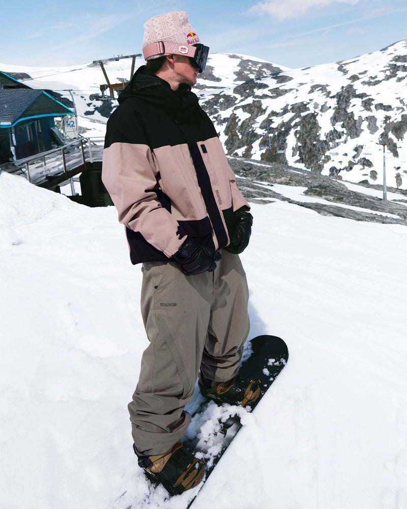 A snowboarder wearing Men's Kleveland Shell Pants, a pink and black jacket, black gloves, and a pink beanie stands on a snowy slope with mountains and a ski lift in the background.