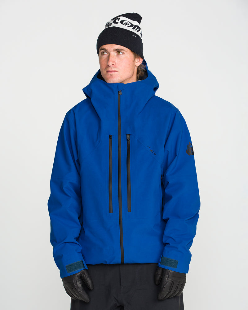 A person wearing the Volcom Men’s Guch 3L Gore-Tex Pro Jacket in blue, featuring a recycled-nylon shell, along with black gloves, black pants, and a black-and-white beanie stands against a plain white background, looking slightly to the side—ready for any backcountry adventure.