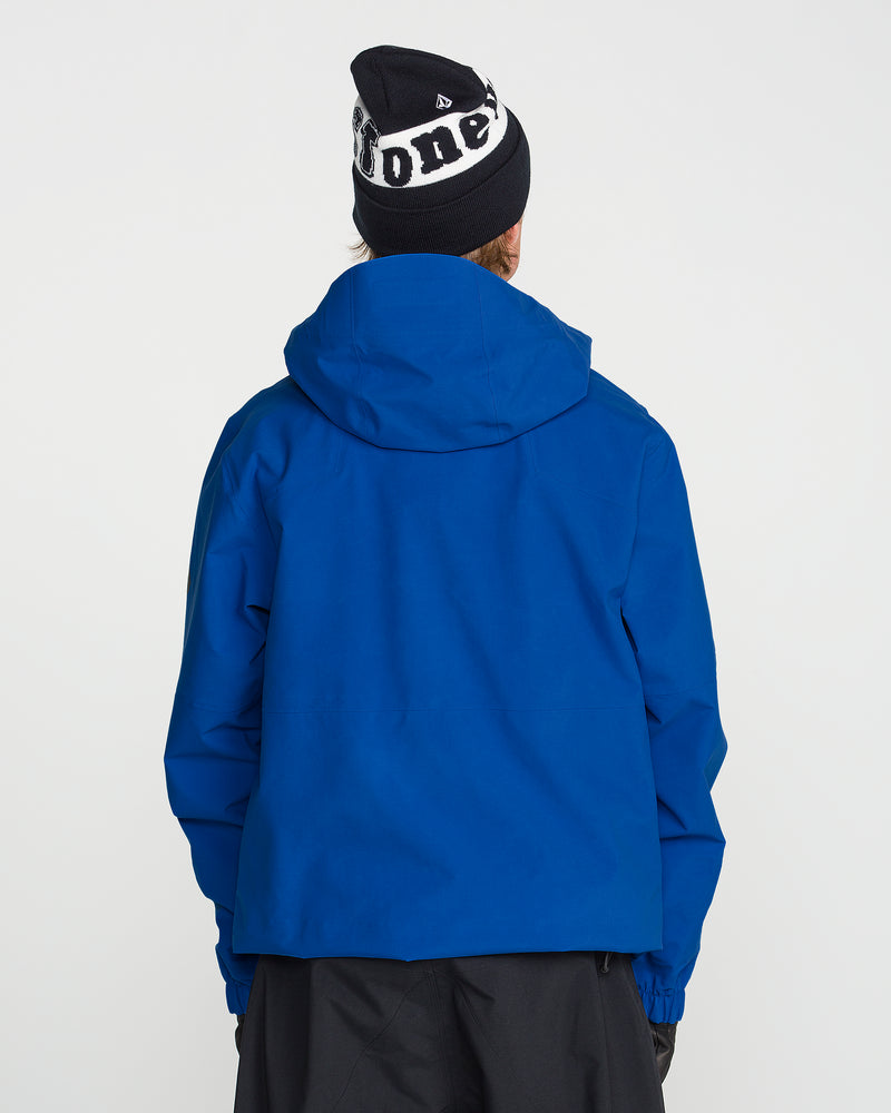 A person stands with their back to the camera against a plain white background, ready for any backcountry adventure, wearing the Volcom Men’s Guch 3L Gore-Tex Pro Jacket featuring a recycled-nylon shell in blue, paired with black pants and a black beanie with white lettering.