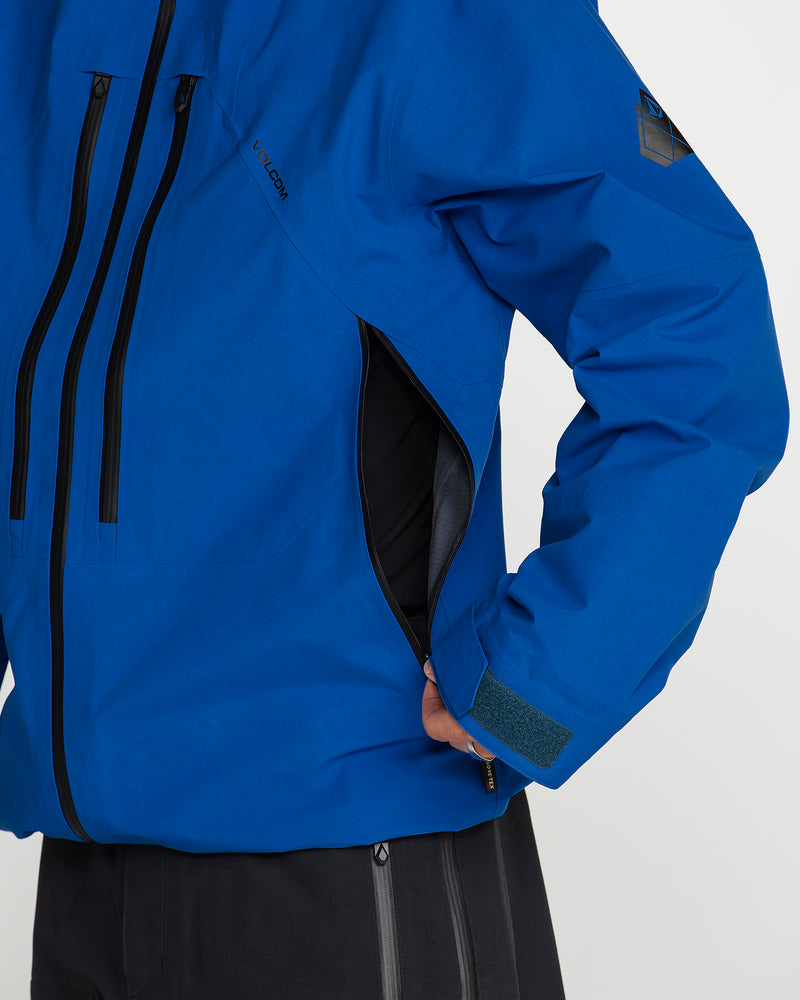 A person sporting the Volcom Men’s Guch 3L Gore-Tex Pro Jacket in bright blue with black zippers and matching black pants stands with a hand on their hip, one recycled-nylon shell side pocket partially unzipped—geared up for any backcountry adventure.