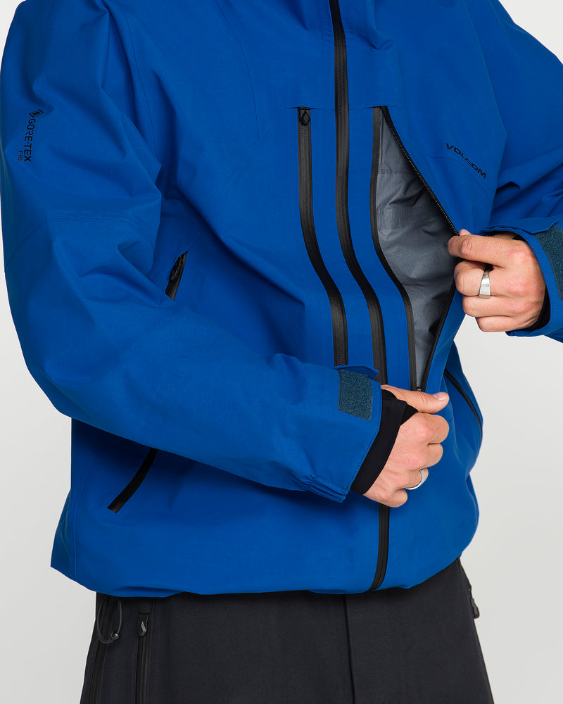A person wearing the Volcom Men’s Guch 3L Gore-Tex Pro Jacket in blue unzips a black chest pocket, revealing the interior. Crafted with a recycled-nylon shell, this premium backcountry essential also features black zippers and adjustable cuffs. Only the torso and hands are visible, emphasizing the jacket’s standout design.
