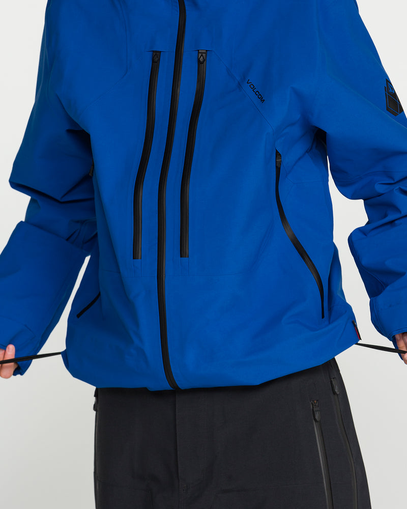 A person wearing the Volcom Men’s Guch 3L Gore-Tex Pro Jacket in bright blue, featuring multiple black zippers and paired with dark pants, adjusts the jacket’s drawstrings. Only the torso, part of the arms, and legs are visible—an ideal look for backcountry adventures.