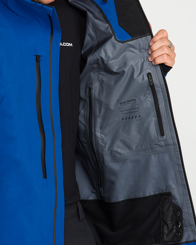 A person opens a blue Volcom Men’s Guch 3L Gore-Tex Pro Jacket to reveal its gray interior lining, featuring a zippered pocket and text detailing. Ideal for backcountry adventures, this jacket boasts a durable recycled-nylon shell.
