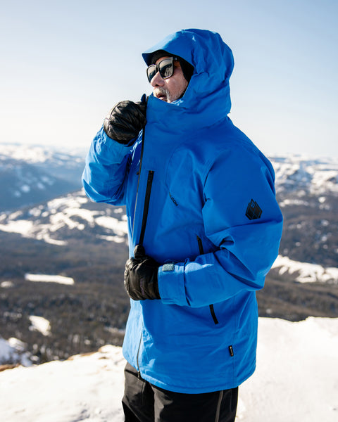 A person wearing the Volcom Men’s Guch 3L Gore-Tex Pro Jacket, featuring a recycled-nylon shell, along with gloves and sunglasses, zips up their coat while standing on a snowy mountain in the backcountry under a clear sky with distant hills in view.