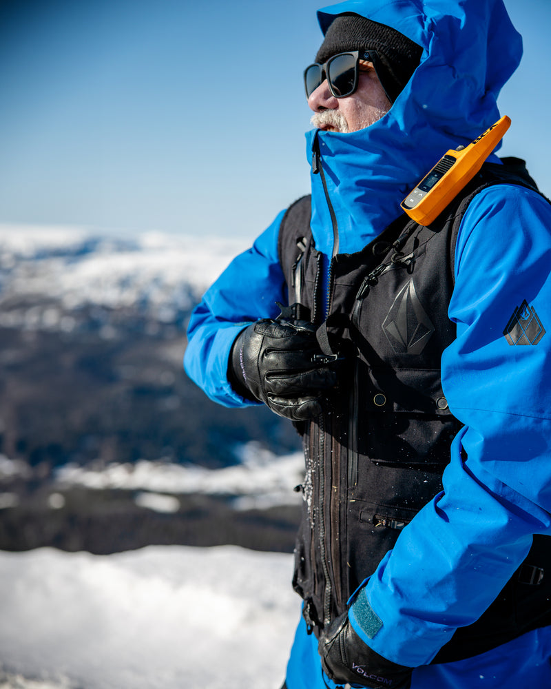 A person wearing the Volcom Men’s Guch 3L Gore-Tex Pro Jacket in blue, paired with black gloves, stands on a snowy mountain prepared for a backcountry adventure. They sport sunglasses and carry a backpack equipped with an orange device, with snowy peaks and a clear blue sky in the background.