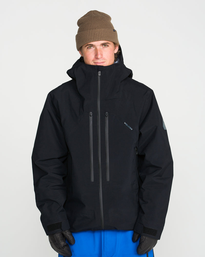A person wearing a brown beanie, black gloves, blue pants, and the Volcom Men’s Guch 3L Gore-Tex Pro Jacket—with its high collar and recycled-nylon shell—stands facing the camera against a plain white background, ready for any backcountry adventure.