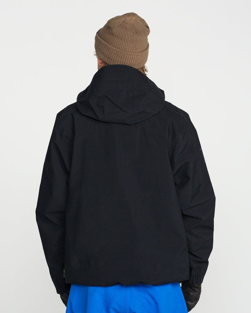 A person stands with their back to the camera, outfitted for any backcountry adventure in a black Volcom Men’s Guch 3L Gore-Tex Pro Jacket featuring a recycled-nylon shell, paired with a brown knit beanie, blue pants, and black gloves—set against a plain white background.