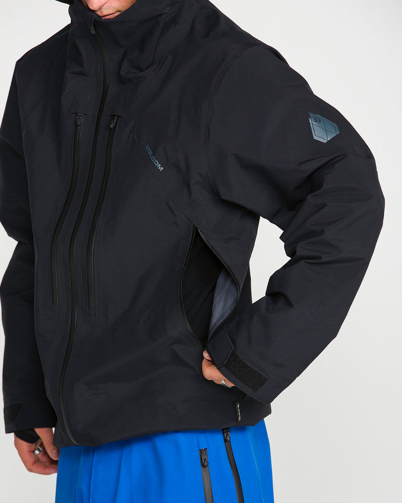A person wearing the Volcom Men’s Guch 3L GORE-TEX Pro Jacket in black with multiple zippers and blue pants unzips an underarm ventilation opening—perfect for backcountry adventures. The photo is cropped to show only the lower face, upper body, and part of the arm.