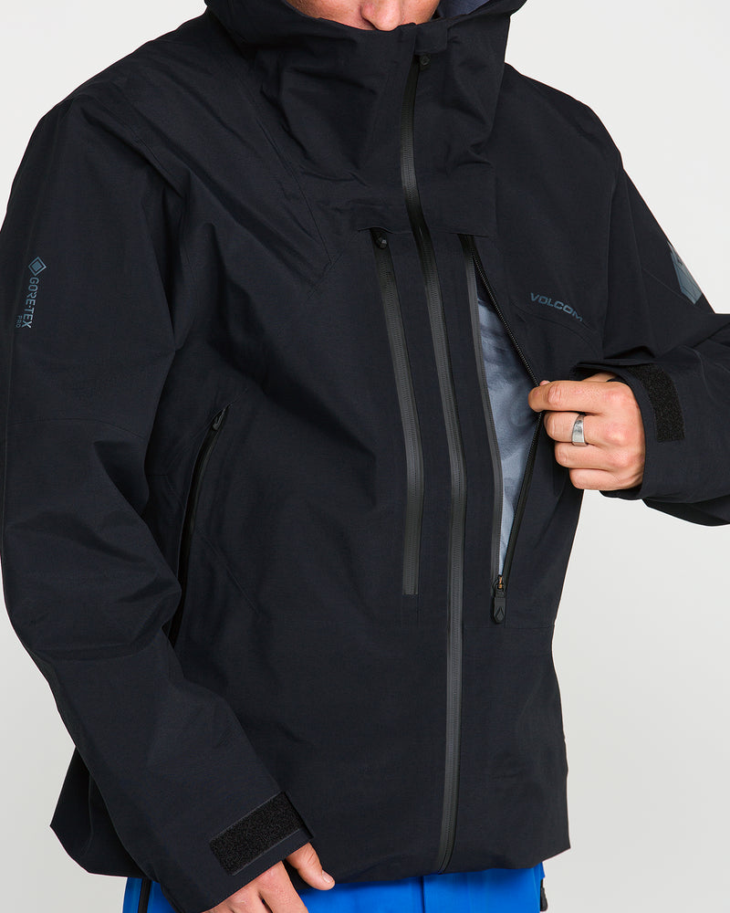 A person wearing the Volcom Men’s Guch 3L Gore-Tex Pro Jacket in black partially unzips the front chest pocket to reveal the interior lining. Engineered for backcountry adventures, this jacket features waterproof zippers, Velcro cuffs, and a durable recycled-nylon shell.