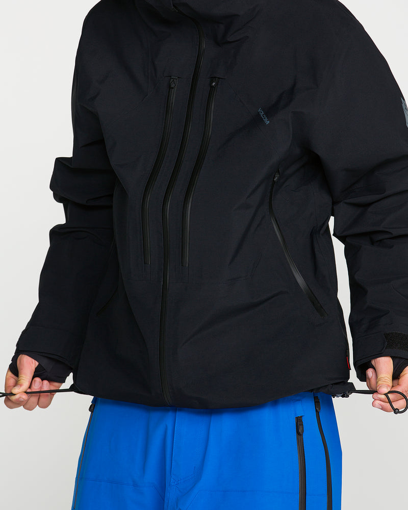 Wearing the Volcom Men’s Guch 3L Gore-Tex Pro Jacket—a black recycled-nylon shell featuring multiple zippers—a person in bright blue pants adjusts the drawcord at the jacket’s hem. Their head is not shown, emphasizing the technical outerwear built by Volcom for backcountry adventures.